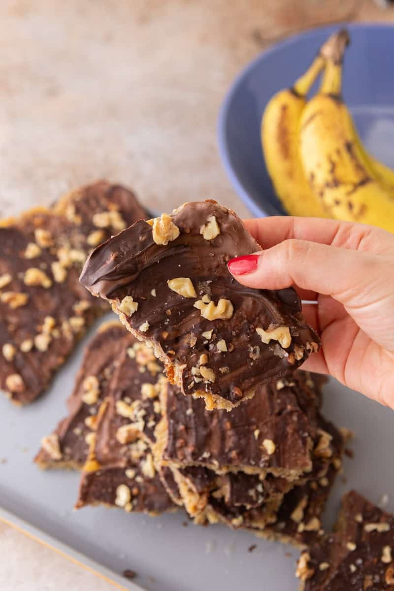 Holding a piece of banana bread Christmas crack topped with chocolate, walnuts, and flaky sea salt.
