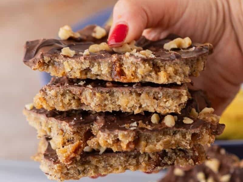 Hand reaching for a stack of banana bread Christmas crack pieces showing layers of chocolate, walnuts, and banana bread.