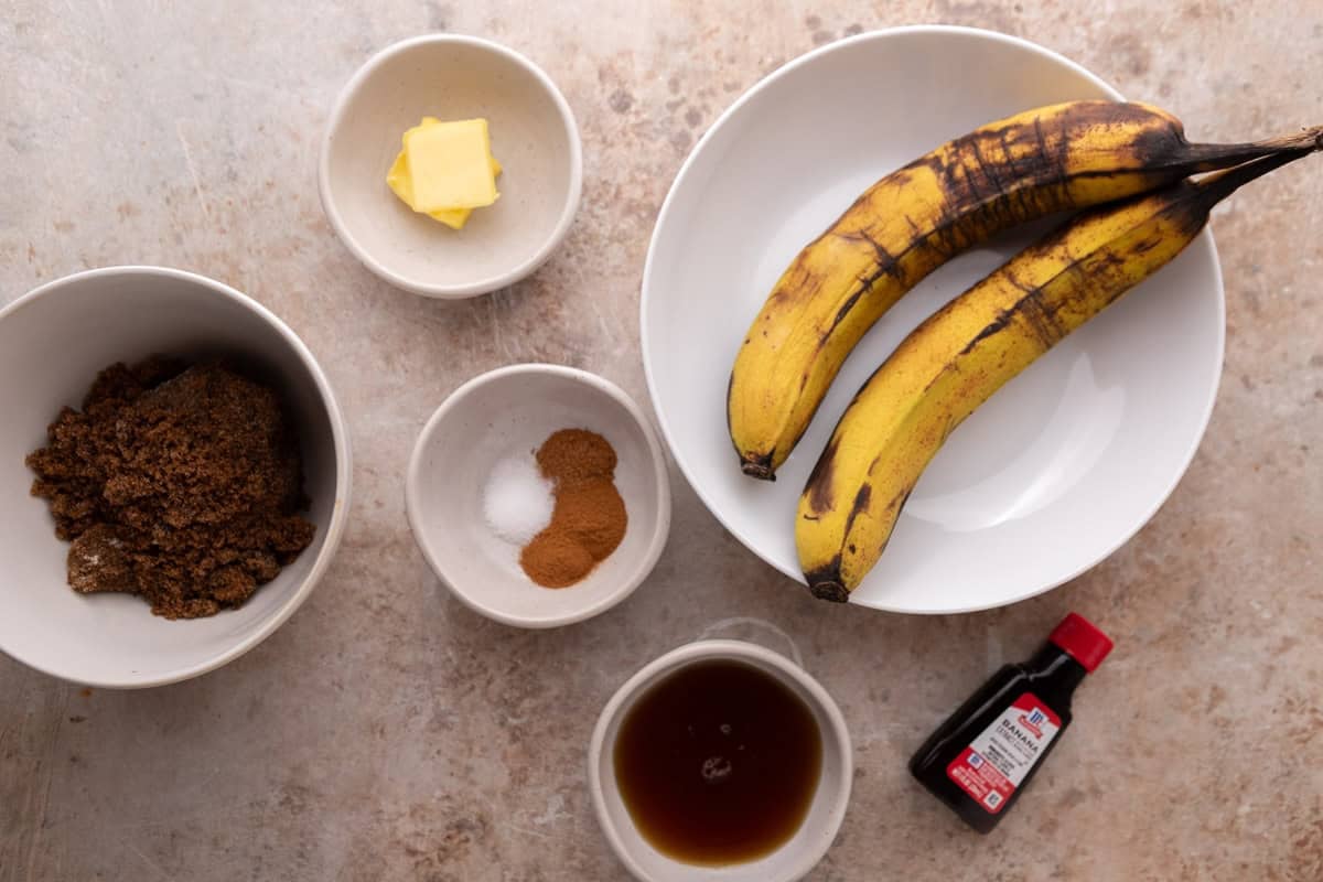 ngredients for banana bread syrup laid out in white bowls including overripe bananas, butter, dark brown sugar, cinnamon, maple syrup, and banana extract