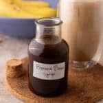 Banana bread syrup in a glass bottle with a handwritten label on a cork trivet next to an iced banana bread latte
