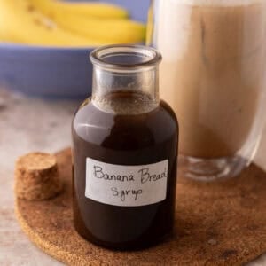 Banana bread syrup in a glass bottle with a handwritten label on a cork trivet next to an iced banana bread latte
