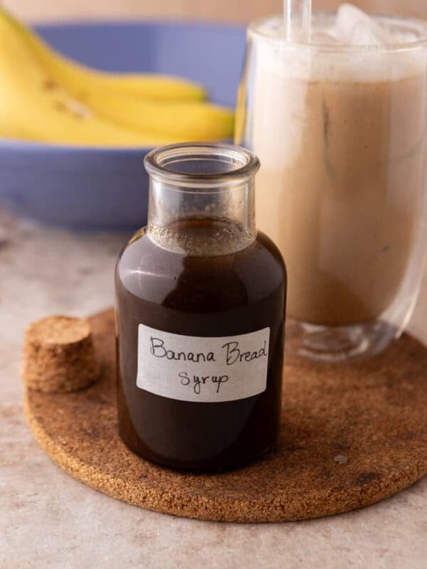 Banana bread syrup in a glass bottle with a handwritten label on a cork trivet next to an iced banana bread latte