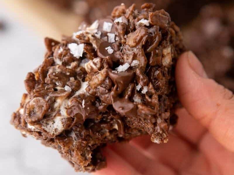 Close up of a chocolate rice krispie treat held in a hand, topped with chocolate chips and flaky sea salt