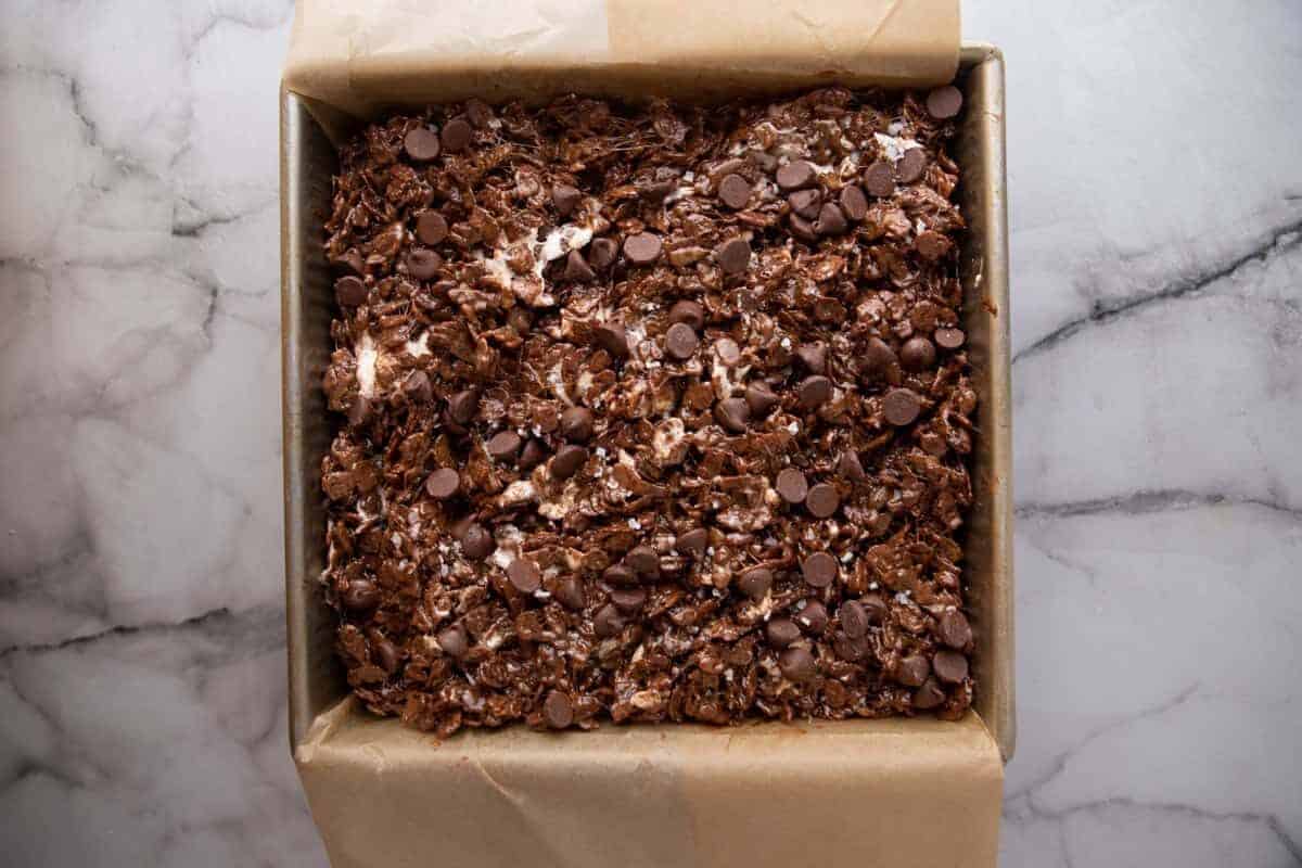 Chocolate rice krispie treat mixture pressed into a parchment-lined square pan and topped with chocolate chips and flaky sea salt