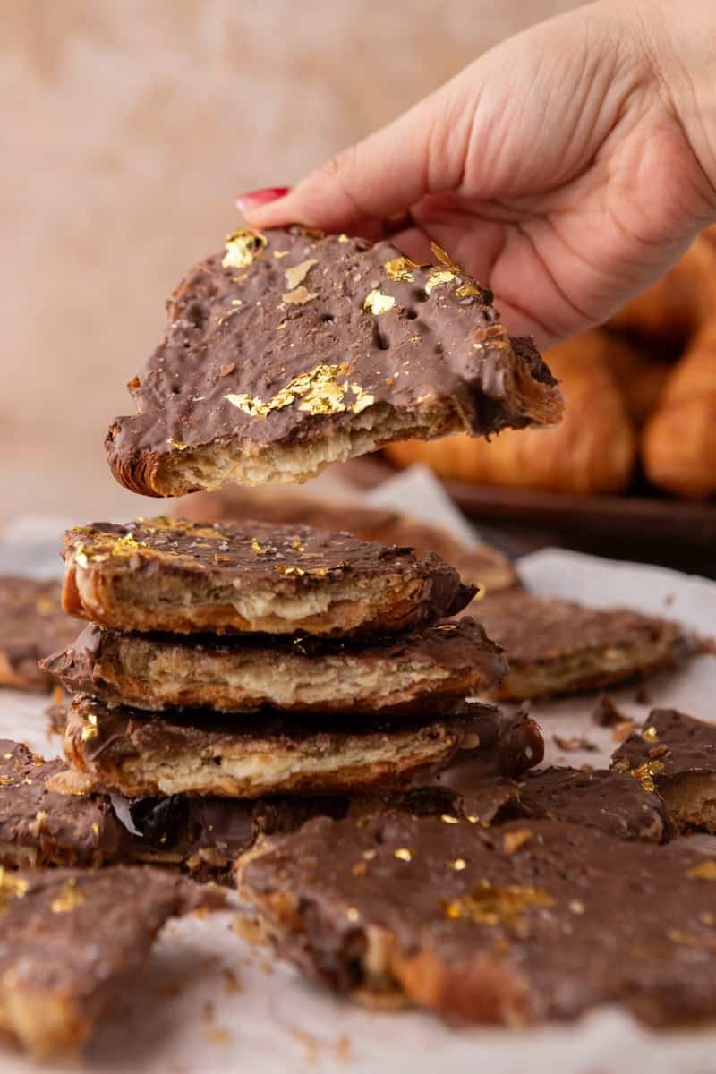 Hand holding a shard of croissant Christmas crack with chocolate and gold leaf above a stack of pieces