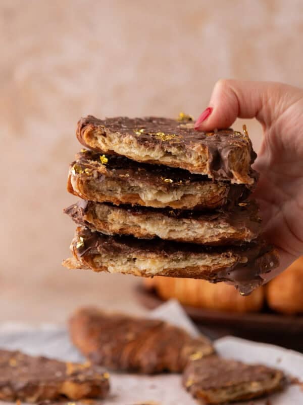 Hand holding a tall stack of croissant Christmas crack pieces with visible flaky layers and gold leaf