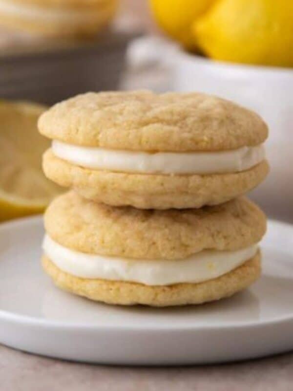 Two lemon sandwich cookies stacked on a small white plate with fresh lemons in the background.