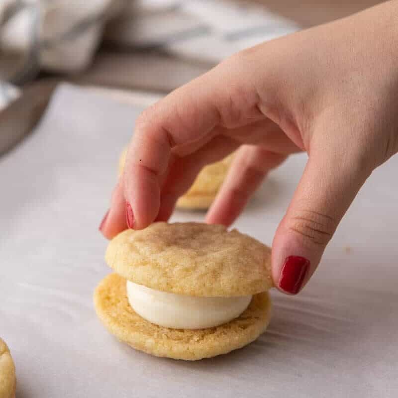 A hand placing the top cookie onto a cream cheese frosted lemon sandwich cookie to assemble it.