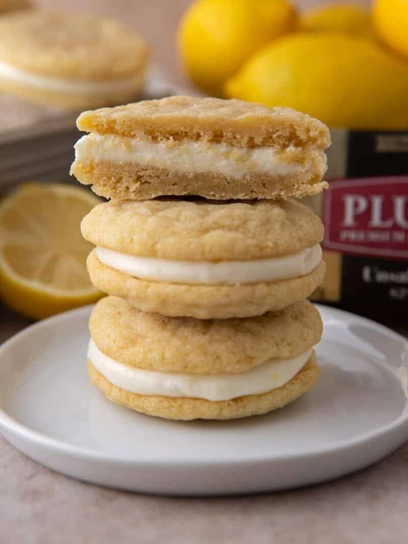 Stack of lemon sandwich cookies with cream cheese filling, top cookie split to show the inside, with lemons and Plugrà butter in the background.