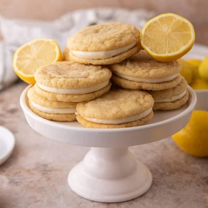 Stack of lemon sandwich cookies with cream cheese frosting filling on a white cake stand with fresh lemon halves.