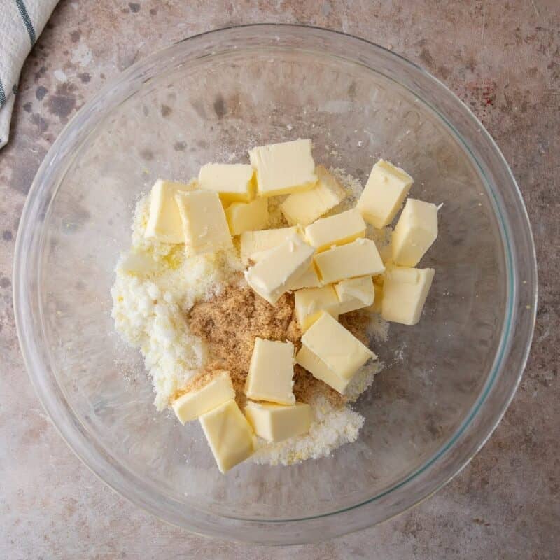 Cubed Plugrà butter added to lemon sugar in a glass bowl, ready to be creamed for cookie dough.