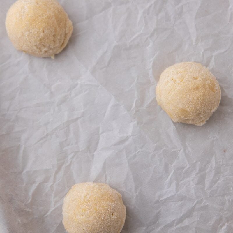 Three scooped lemon cookie dough balls rolled in sugar on a parchment-lined baking sheet.