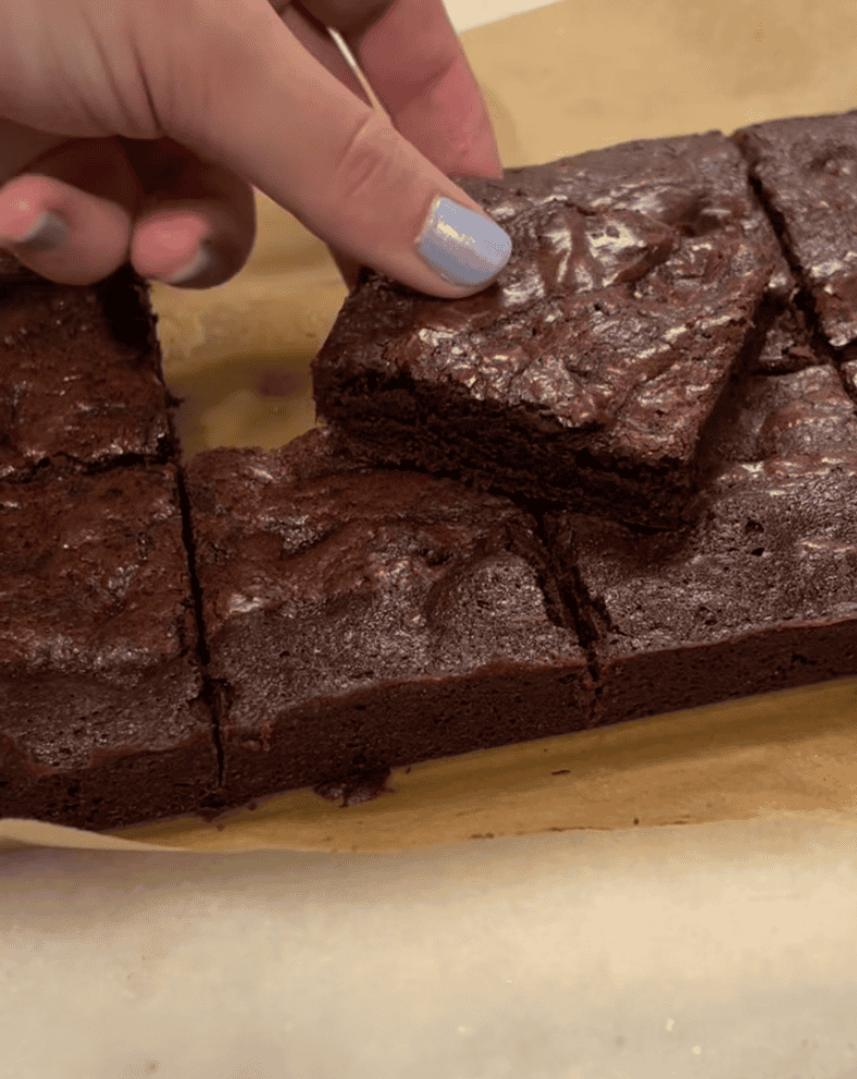 Hand reaching for a fudgy chocolate brownie square cut on parchment paper for Loaf BakeClub