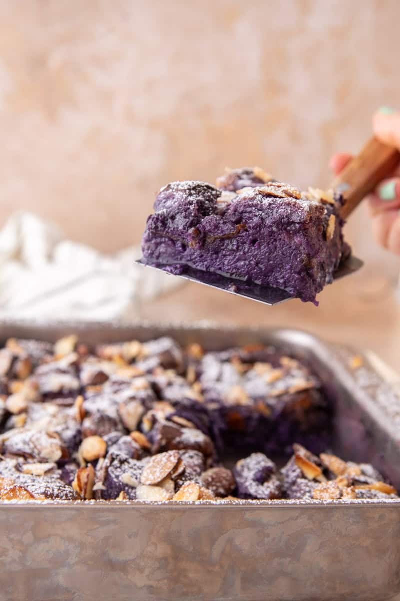 A spatula lifting a slice of ube French toast bake out of the pan revealing the custardy purple interior