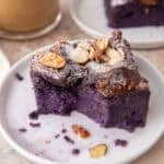 Ube French toast bake slice on a small white plate dusted with powdered sugar and toasted almonds with coffee in the background
