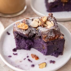Ube French toast bake slice on a small white plate dusted with powdered sugar and toasted almonds with coffee in the background