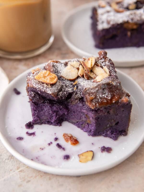 Ube French toast bake slice on a small white plate dusted with powdered sugar and toasted almonds with coffee in the background