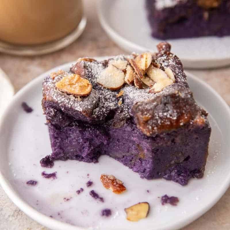 Ube French toast bake slice on a small white plate dusted with powdered sugar and toasted almonds with coffee in the background