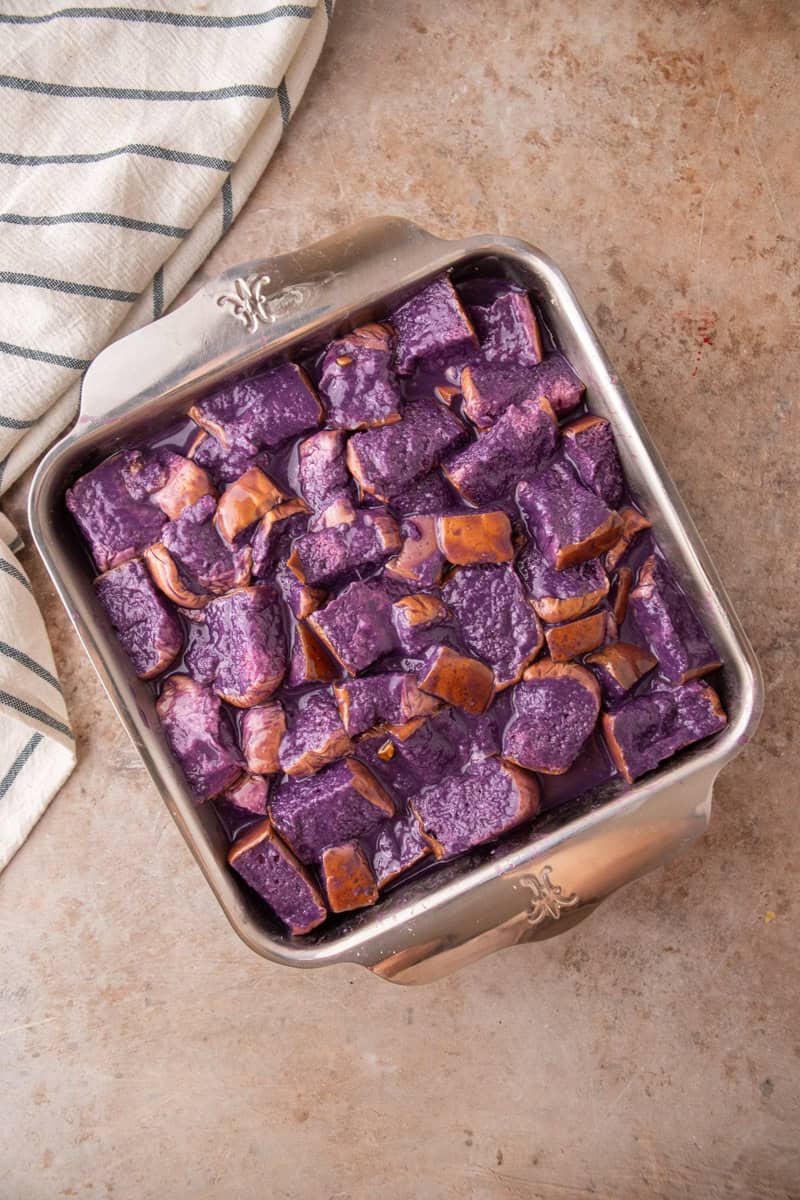 Cubed Hawaiian rolls soaking in purple coconut milk custard in a square metal baking dish