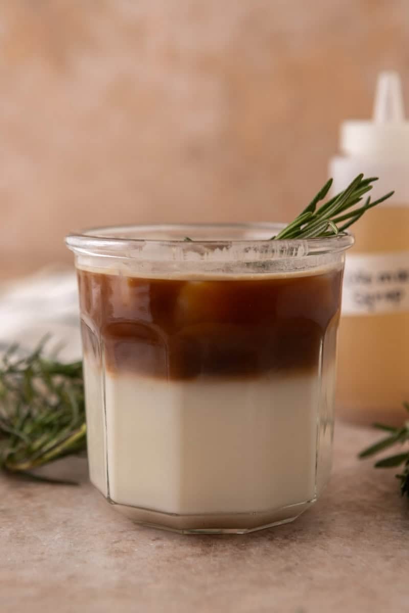 Iced rosemary latte with milk on the bottom and a layer of espresso on top, garnished with a rosemary sprig.