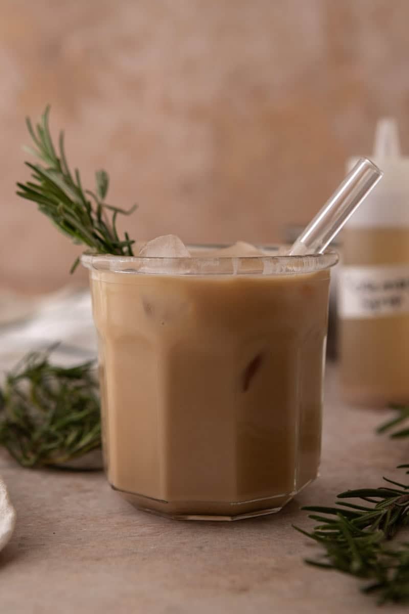 Stirred iced rosemary latte in a glass with a straw and rosemary sprigs beside it.