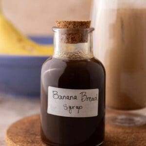 Banana bread syrup in a glass bottle with a handwritten label on a cork trivet next to an iced banana bread latte.