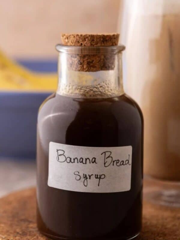 Banana bread syrup in a glass bottle with a handwritten label on a cork trivet next to an iced banana bread latte.