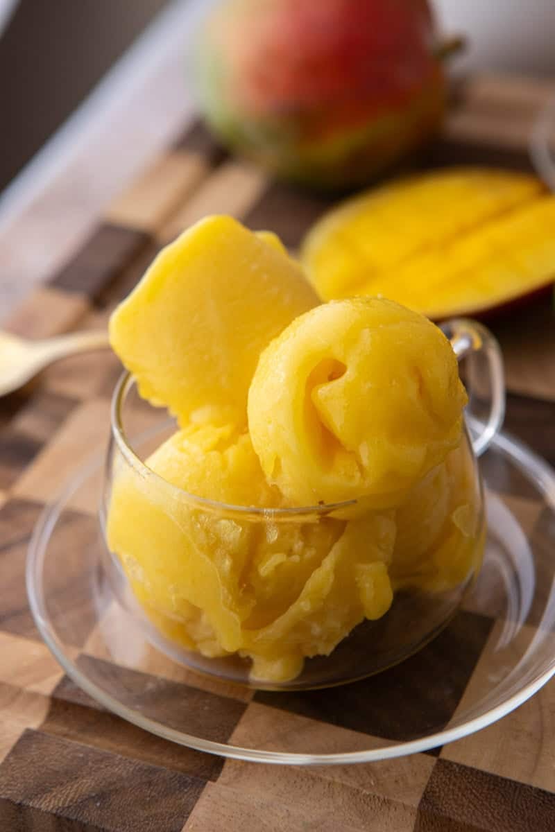 Scoops of velvety mango sorbet in a clear glass cup with fresh mango in the background