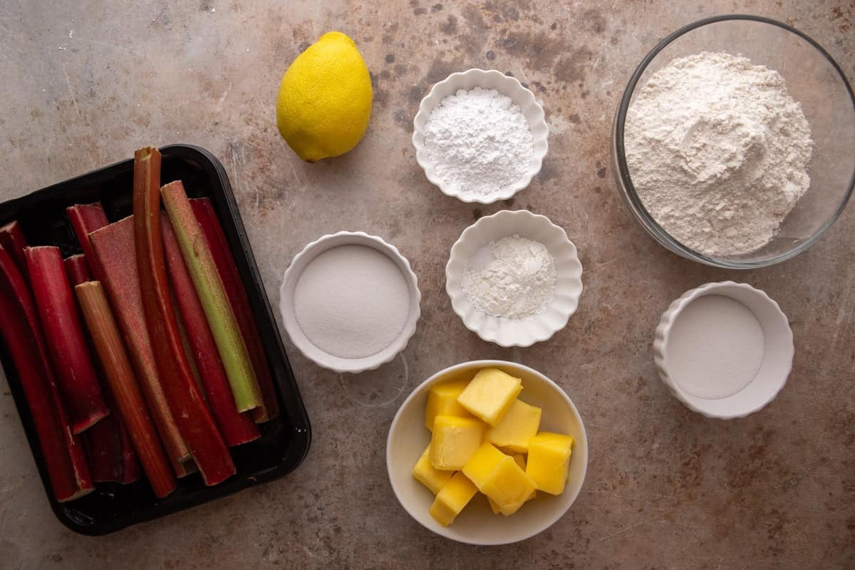 Overhead flat lay of rhubarb crumble bar ingredients including fresh rhubarb stalks, cubed butter, flour, powdered sugar, granulated sugar, cornstarch, and a lemon