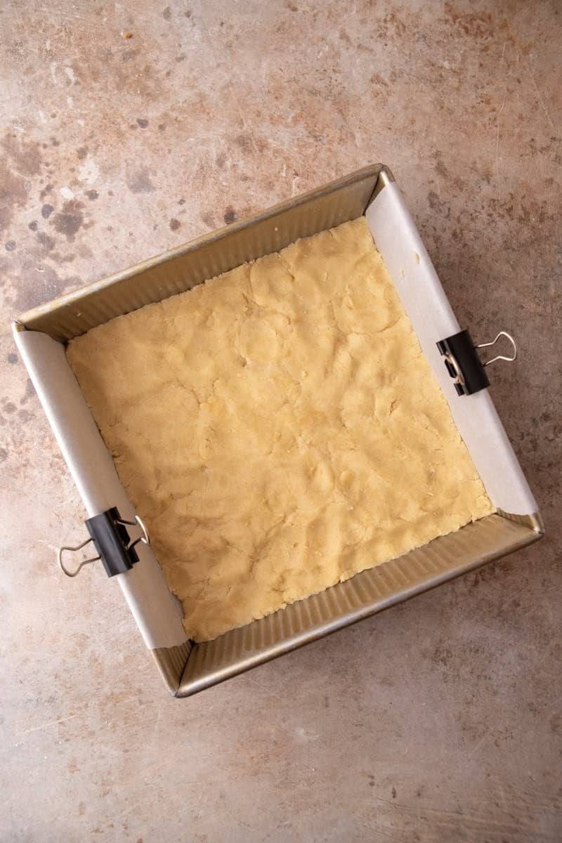 Shortbread dough pressed evenly into the bottom of a parchment-lined 8x8 gold baking pan before par-baking