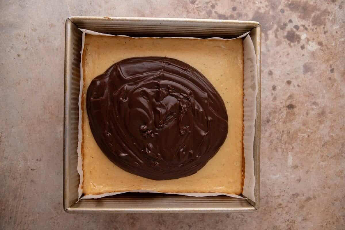 Chocolate ganache being spread over the baked marshmallow cheesecake layer for s'mores cheesecake bars