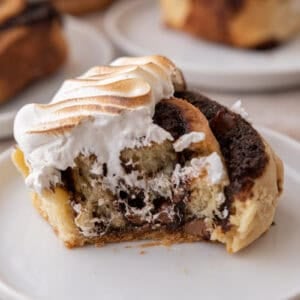 S'mores cinnamon roll topped with toasted meringue on a white plate with a bite taken out