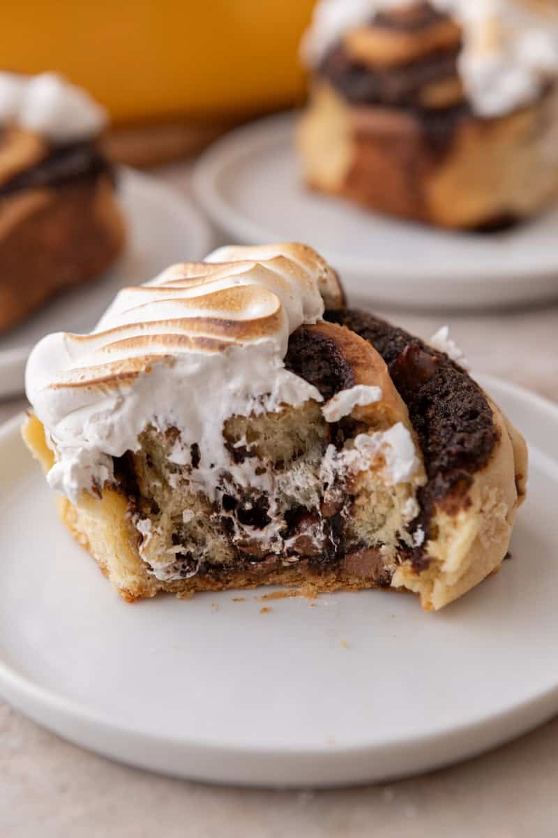 S'mores cinnamon roll topped with toasted meringue on a white plate with a bite taken out