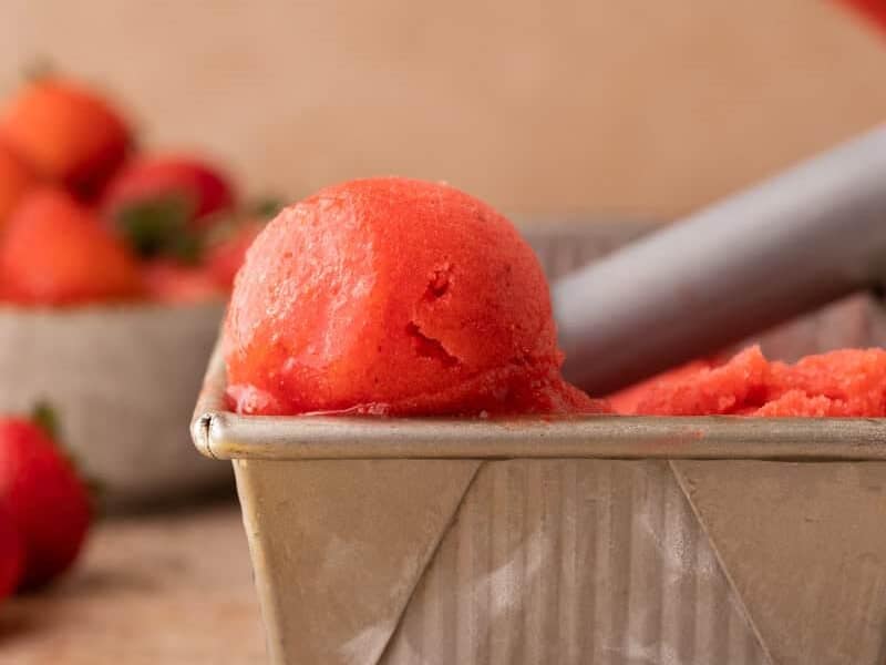 Scoop of bright red strawberry sorbet in a metal loaf pan with fresh strawberries.