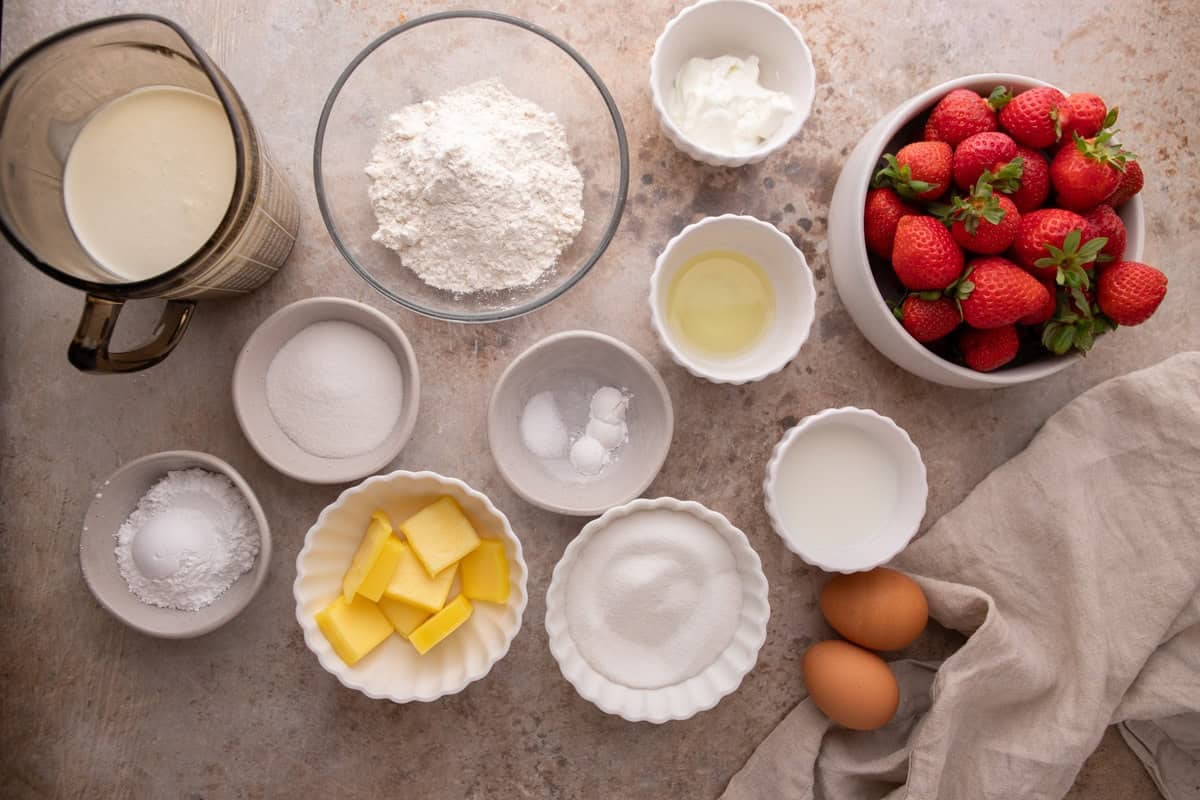 Ingredients for strawberry shortcake trifle cups in bowls including flour, butter, sugar, sour cream, eggs, heavy cream, and fresh strawberries