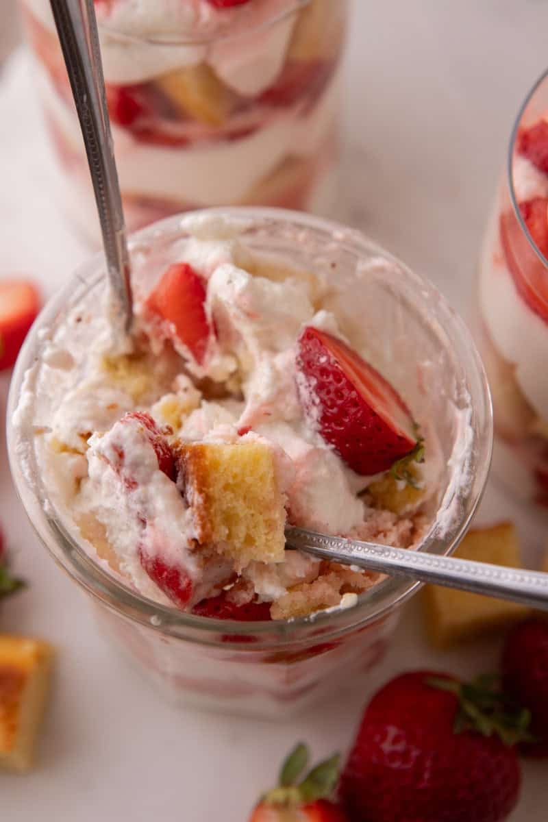 Overhead view of a spoon digging into a strawberry shortcake trifle cup with visible cake cubes and berries