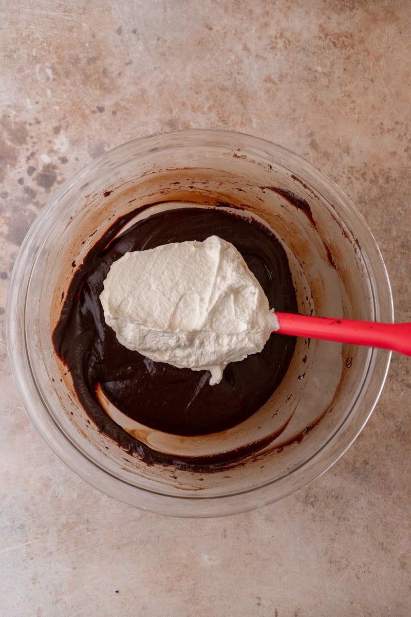 First addition of whipped cream being folded into the dark chocolate mixture with a red rubber spatula