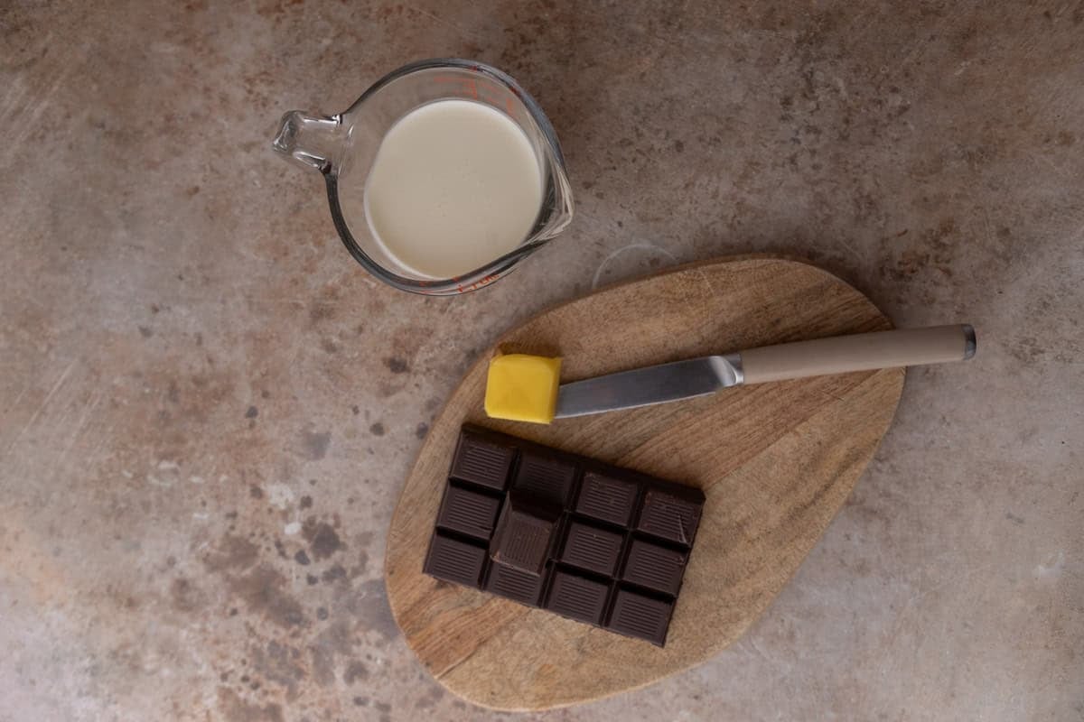 Ingredients for the chocolate ganache including heavy cream in a glass measuring cup, a dark chocolate bar, and butter on a wooden cutting board