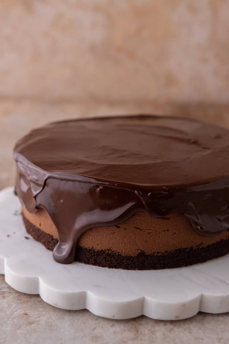 Chocolate ganache being poured over the chocolate mousse cake, dripping down the sides, on a white scalloped marble cake stand