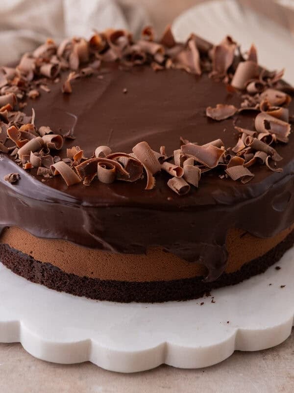 Chocolate mousse cake with glossy ganache and chocolate curls on a white scalloped marble cake stand.