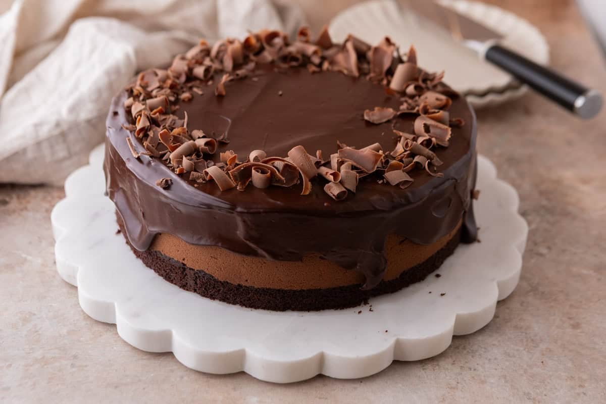 Chocolate mousse cake on a white scalloped marble cake stand, topped with chocolate ganache and chocolate curls, with a cake server in the background