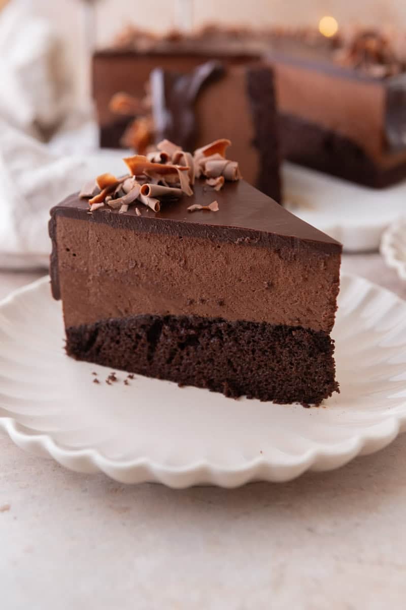 A slice of chocolate mousse cake on a white scalloped plate showing three distinct layers of chocolate sponge cake, chocolate mousse, and chocolate ganache topped with chocolate curls