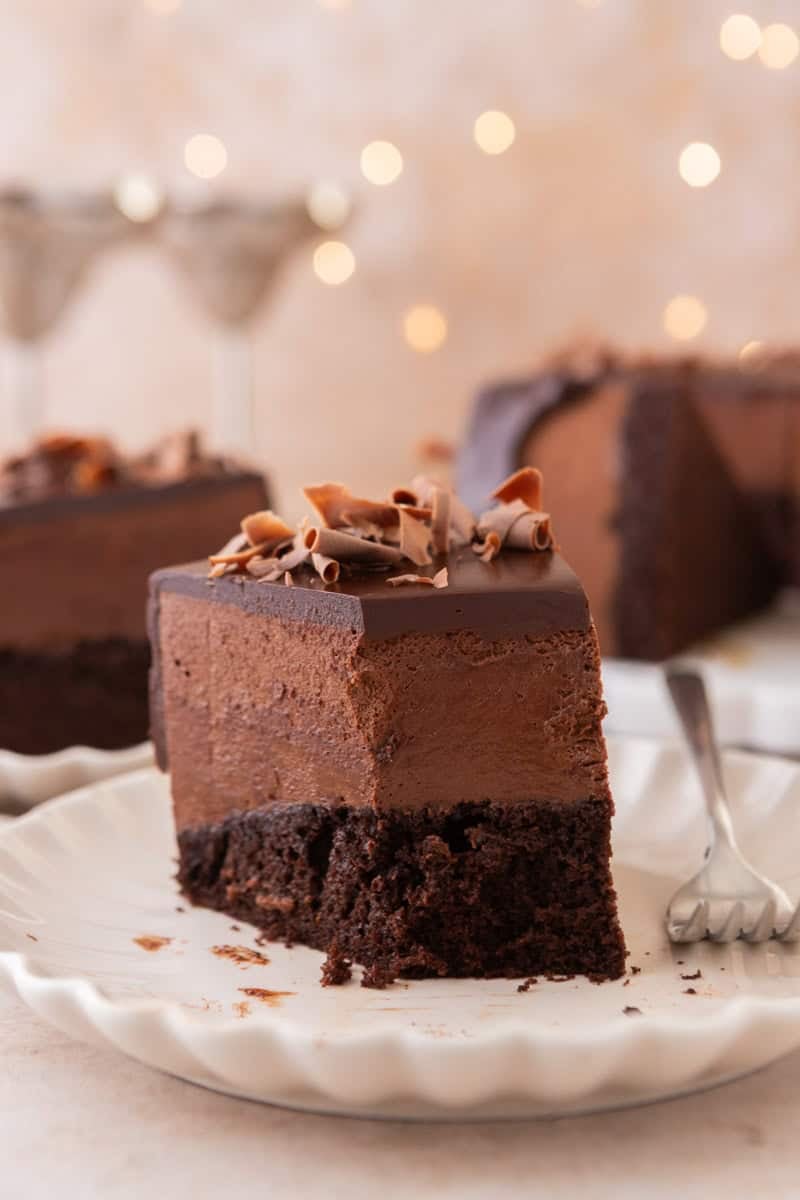 A slice of chocolate mousse cake on a plate with a fork, with the rest of the cake and soft bokeh lights in the background