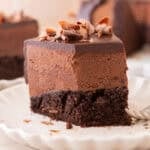 A tall slice of chocolate mousse cake on a white scalloped plate showing the moist chocolate sponge base, thick airy chocolate mousse layer, and glossy ganache topped with chocolate curls, with bokeh lights in the background