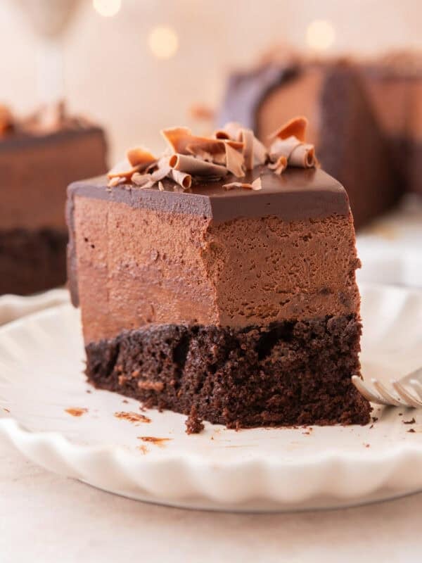 A tall slice of chocolate mousse cake on a white scalloped plate showing the moist chocolate sponge base, thick airy chocolate mousse layer, and glossy ganache topped with chocolate curls, with bokeh lights in the background