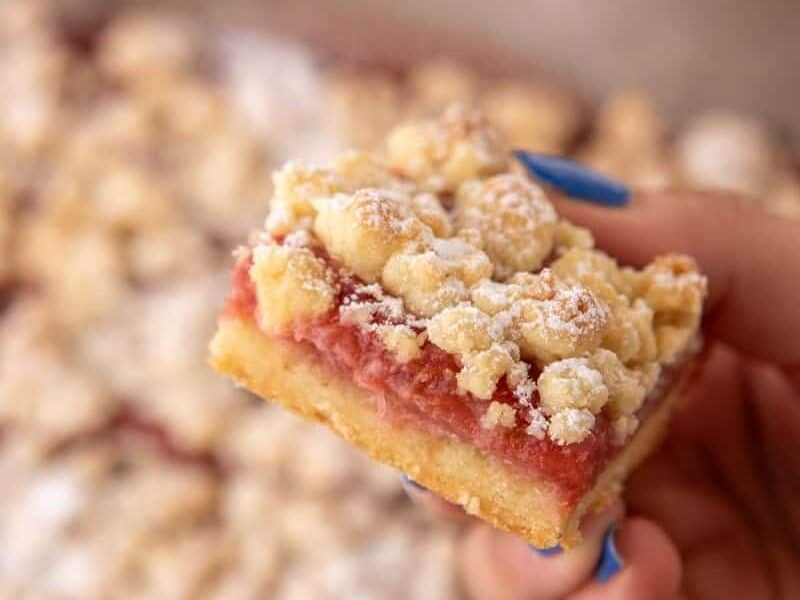 A hand with blue nails holding a single rhubarb crumble bar close up, showing the jammy filling and buttery crumble layers