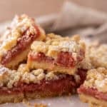 Stack of rhubarb crumble bars on parchment paper showing layers of golden shortbread base, pink jammy rhubarb filling, and crumble topping
