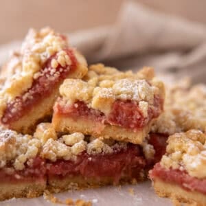 Stack of rhubarb crumble bars on parchment paper showing layers of golden shortbread base, pink jammy rhubarb filling, and crumble topping