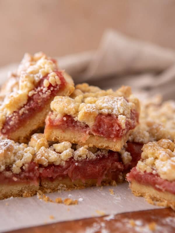 Stack of rhubarb crumble bars on parchment paper showing layers of golden shortbread base, pink jammy rhubarb filling, and crumble topping