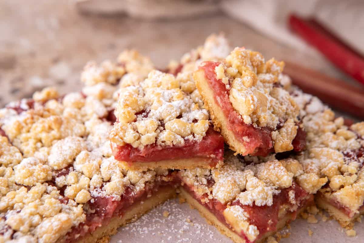 Rhubarb crumble bars stacked on a pink surface dusted with powdered sugar, with fresh rhubarb stalks in the background